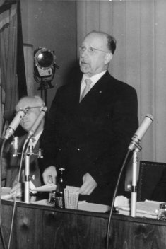 Walter Ulbricht steht in der Bildmitte und spricht in das vor ihm stehende Mikrofon. Rechts und links neben ihm sitzen drei Männer, die in Unterlagen blättern. Im linken Bildhintergrund sind DDR-Fahnen drapiert.