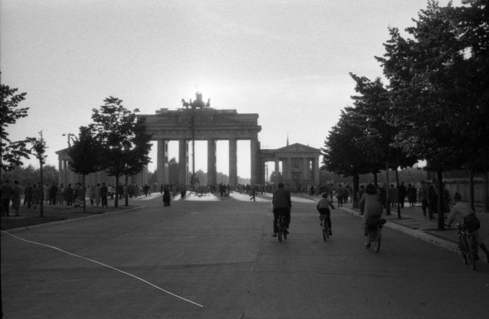 Das Brandenburger von der Straße Unter den Linden in Richtung Tiergarten fotografiert. Dutzende von Menschen sind zu Fuß oder auf dem Fahrrad unterwegs.