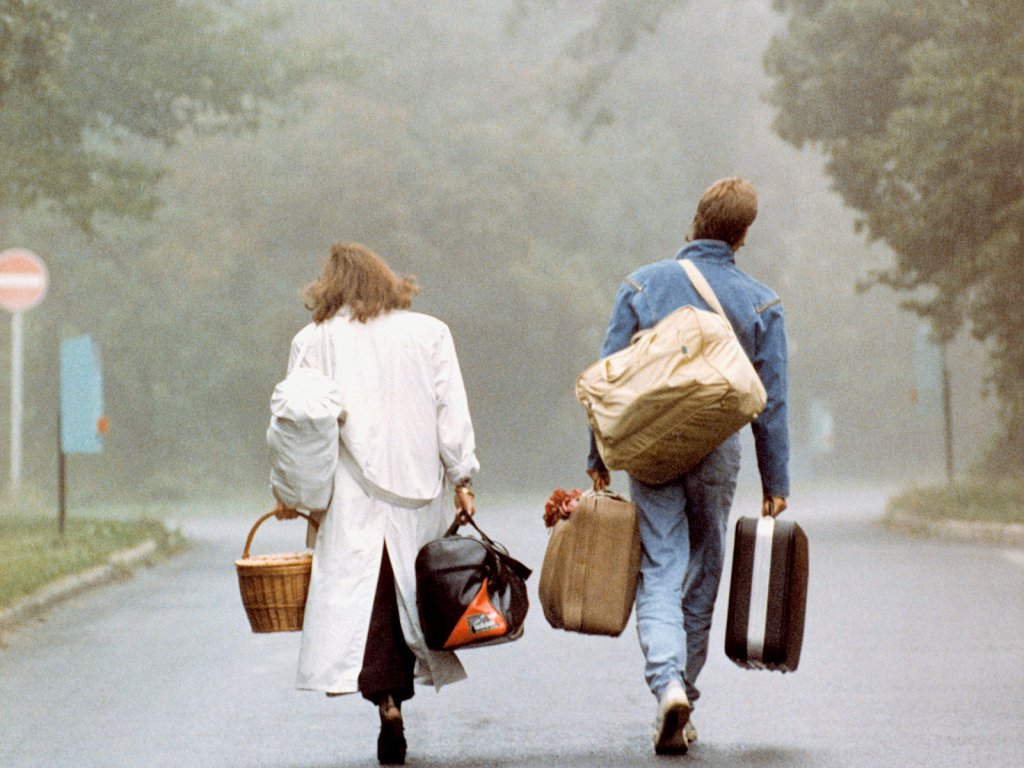 Ein junges Paar aus der DDR kommt wenige Tage vor der Öffnung der ungarisch-österreichischen Grenze im Flüchtlingslager Csilleborc bei Budapest an, 4. September 1989. Eine Frau und ein Mann sind von hinten zu sehen, wie sie mit viel Gepäck im Nebel auf eine Straßengabelung zugehen.