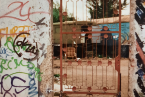 Large section of the Berlin Wall covered in graffiti. In the middle of the wall is a red iron gate allowing a view through the wall and onto two caravans in the background.