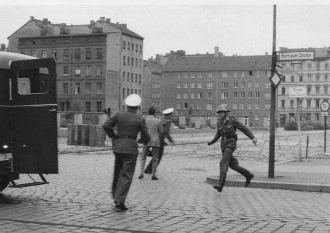 „Sprung in die Freiheit“, Bernauer Straße, 15. August 1961: Der DDR-Grenzpolizist Conrad Schumann rennt nach seinem Sprung zu einem Streifenwagen der West-Berliner Polizei. Zwei Beamte bergen seine Waffe „Sprung in die Freiheit“, Bernauer Straße, 15. August 1961: Der DDR-Grenzpolizist Conrad Schumann rennt nach seinem Sprung zu einem Streifenwagen der West-Berliner Polizei. Zwei Beamte bergen seine Waffe