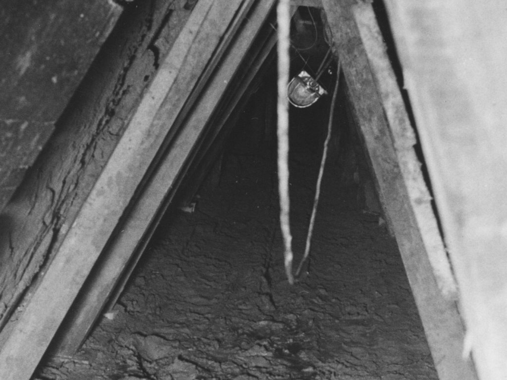 Der „Tunnel 29" – ein logistisches Meisterstück, durch den 29 Personen im September 1962 in den Westen fliehen Das Bild ist frontal in die dreieckige Tunnelöffnung aufgenommen. Der Boden ist schlammig, an den Stützbalken hängt eine Lampe.