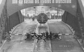 Vogelperspektive auf die Bösebrücke, die mit Panzersperren und Schlagbäumen gesperrt ist. Mittig stehen Soldaten. Die Brücke ist beschriftet mit: DDR – die Bastion des Friedens in Deutschland. Unter dem Schriftzug hängt das DDR-Staatswappen.