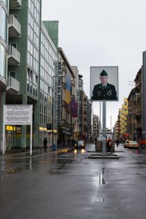 Checkpoint Charlie on a rainy day. On the left is the sector sign with the inscription: “Your are entering the american sector. Carrying weapons off dutyforbidden. Obey traffic rules.” In the middle of the frame is the Mauermuseum. On the right is the control barracks with sandbags. In front of it is the lightbox rising above the street. It shows a larger than scale photo of an american soldier.