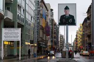 Checkpoint Charlie on a rainy day. On the left is the sector sign with the inscription: “Your are entering the american sector. Carrying weapons off dutyforbidden. Obey traffic rules.” In the middle of the frame is the Mauermuseum. On the right is the control barracks with sandbags. In front of it is the lightbox rising above the street. It shows a larger than scale photo of an american soldier.