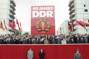 Auf einer rot verkleideten Tribüne stehen dutzende Menschen, die meisten von ihnen sind Männer in Anzügen, Hüten und Mänteln. Hinter ihnen steht ein riesiges rotes Plakat mit dem DDR-Staatsemblem und der weißen Aufschrift: 40 Jahre DDR. Links und rechts davon wehen DDR-Flaggen und rote Fahnen. Im Hintergrund sind die Wohnblöcke der Berliner Karl-Marx-Allee erkennbar.