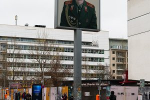 Checkpoint Charlie on a rainy day. In the center of the photo is the lightbox rising above the street. It shows a larger than scale photo of a russian soldier. In the background is an intersection and the Checkpoint Charlie BlackBox.