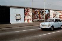 Portion of the East Side Gallery with the paintings from Hunter and Russell in the background. Closer to the camera is the painting of a Trabi breaking through the wall. Above the Trabi is written “Test the Best.” On the street in the foreground is a passing Trabi.