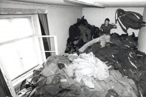 Ein Mann sitzt in einem Zimmer auf einem Berg von Uniformen und wirft von hinten eine Jacke durch den Raum.