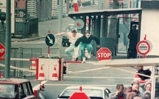 Im Bildzentrum sind die beiden Männer beim Sprung in der Luft über dem Schlagbaum. Die Farbaufnahme wirkt chaotisch durch viele Verkehrsschilder und Absperrungen. Im Vordergrund stehen zwei Autos und eine Menschengruppe, im Hintergrund ist ein Grenzbeamter in der Tür des Grenzgebäudes zu erkennen.