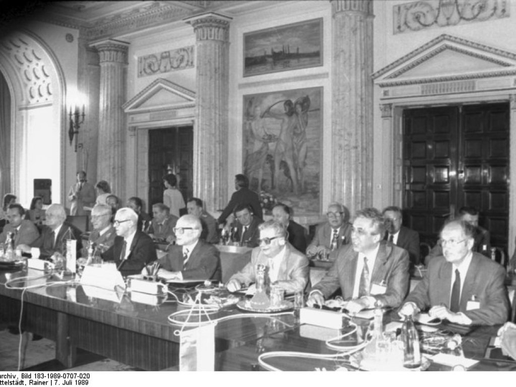 In einem großen Saal mit imposanten Marmorsäulen, Fresken, Stuck und Kassettentüren sitzen mehrere Männer in Anzügen an langen Tischreihen nebeneinander.