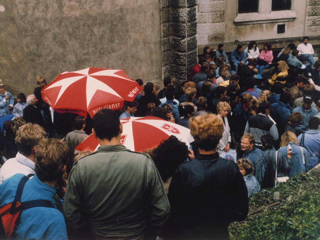 Eine Menschenmenge steht vor einer Hauswand eng aneinandergedrängt, die meisten mit dem Rücken zur Kamera. Aus der Menge ragen zwei Sonnenschirme mit dem roten Malteserkreuz und dem Schriftzug Malteser Hilfsdienst heraus.