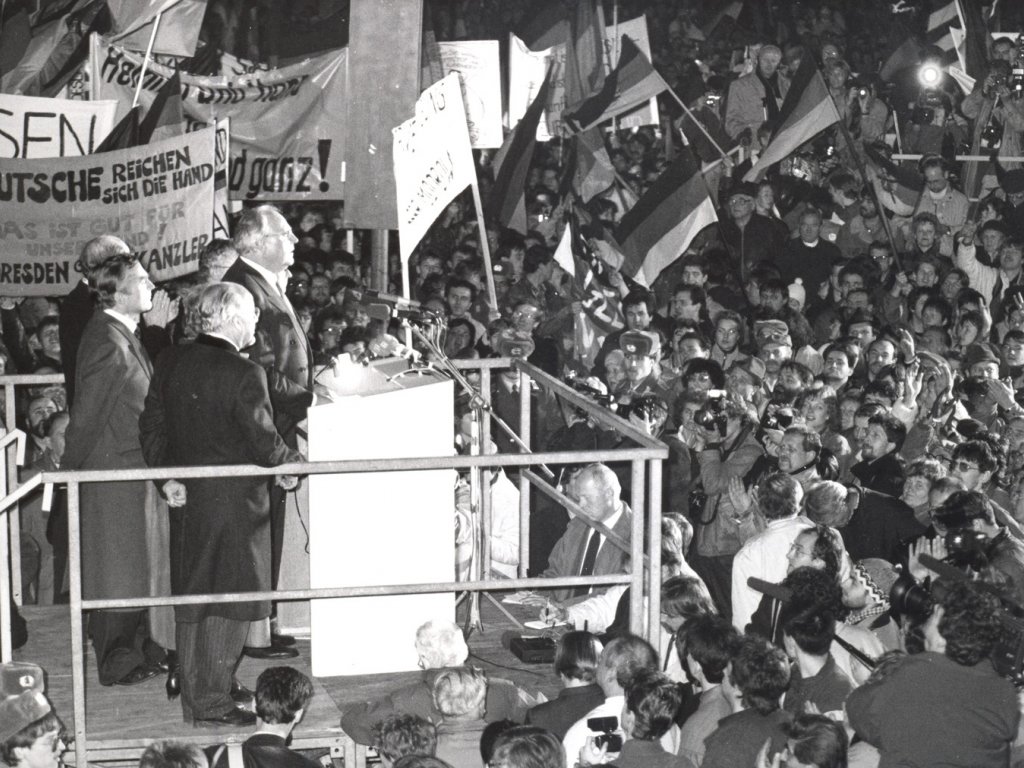 Helmut Kohl steht auf einem Podest vor einem Rednerpult mit mehreren Mikrofonen. Fünf Männer stehen hinter und neben ihm. Rund um das Podest stehen Menschenmassen mit Deutschlandfahnen und Transparenten. Journalisten machen Film- und Fotoaufnahmen.