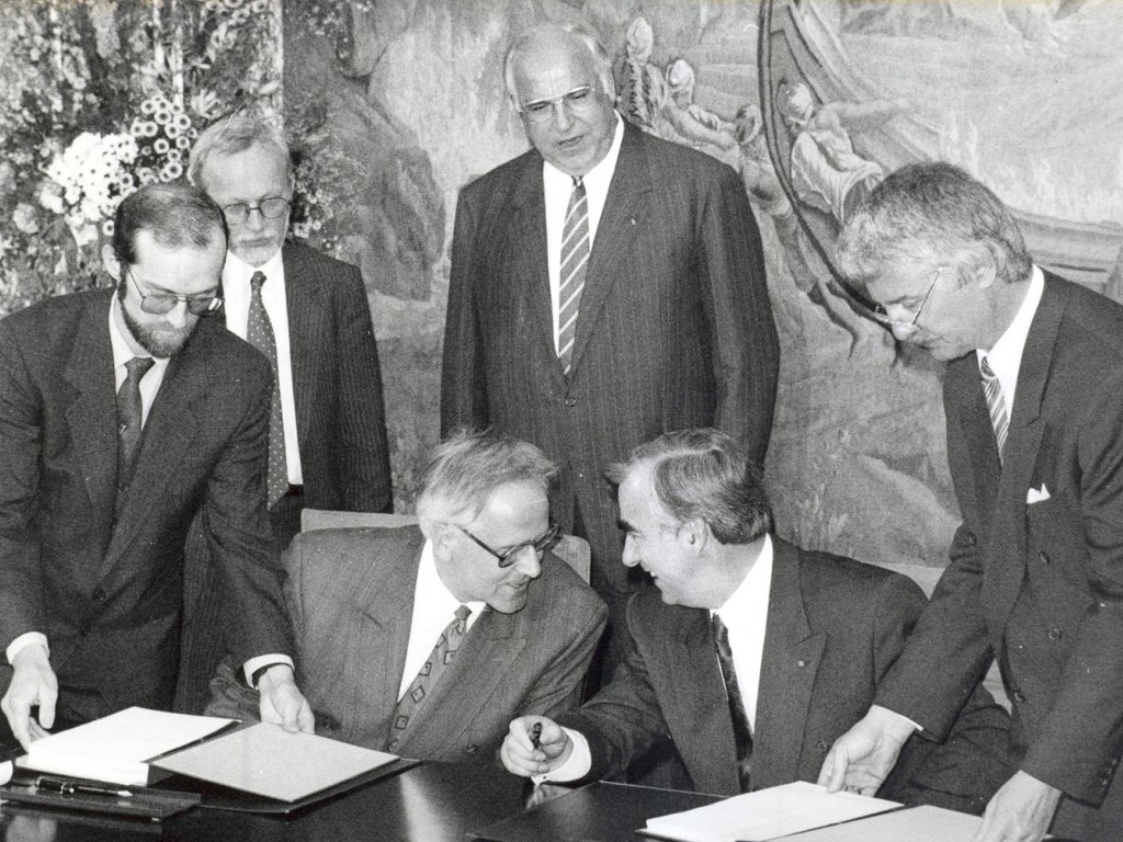 Im Bonner Palais Schaumburg wird der Staatsvertrag zwischen der BRD und der DDR unterzeichnet, 18. Mai 1990. Walter Romberg und Theo Waigel sitzen nebeneinander an einem Tisch, neigen ihre Köpfe zueinander und unterhalten sich lachend. Zwei Männer legen ihnen von rechts und von links den Staatsvertrag zur Unterschrift vor. Hinter Romberg und Waigel stehen links Lothar de Maizière und rechts Helmut Kohl, den Blick auf sie gerichtet.