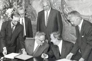Walter Romberg und Theo Waigel sitzen nebeneinander an einem Tisch, neigen ihre Köpfe zueinander und unterhalten sich lachend. Zwei Männer legen ihnen von rechts und von links den Staatsvertrag zur Unterschrift vor. Hinter Romberg und Waigel stehen links Lothar de Maizière und rechts Helmut Kohl, den Blick auf sie gerichtet.