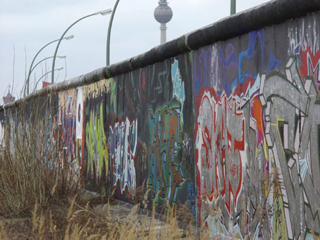 Mittig steht die Mauer, mit Graffiti besprüht. Dahinter sind eine Reihe Bogenleuchten und der Fernsehturm zu erkennen.