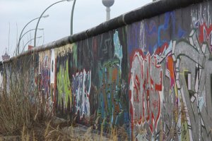 Mittig steht die Mauer, mit Graffiti besprüht. Dahinter sind eine Reihe Bogenleuchten und der Fernsehturm zu erkennen.