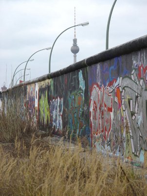 Mittig steht die Mauer, mit Graffiti besprüht. Dahinter sind eine Reihe Bogenleuchten und der Fernsehturm zu erkennen.