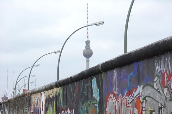 Mittig steht die Mauer, mit Graffiti besprüht. Dahinter sind eine Reihe Bogenleuchten und der Fernsehturm zu erkennen.