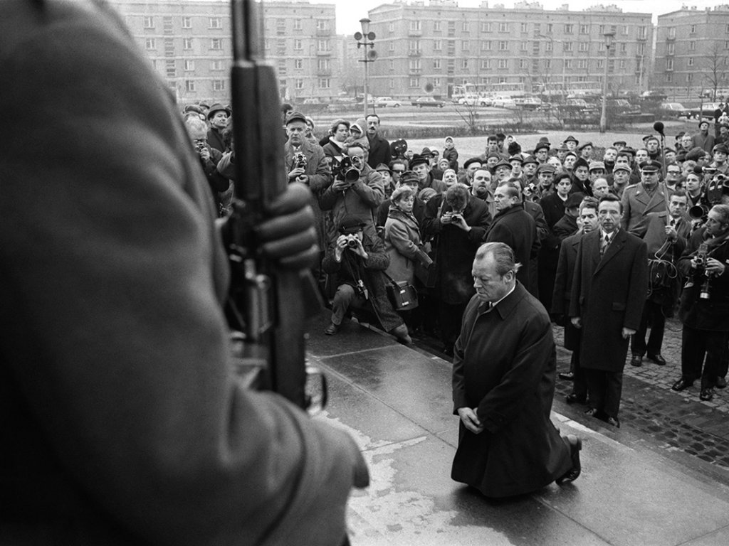 Links im Bildvordergrund ist die Schulter eines Soldaten mit Gewehr zu erkennen. Im Bildzentrum kniet Willy Brandt, er hält die Hände verschränkt vor sich und den Blick nach unten gerichtet. Hinter ihm befindet sich eine Menschenmenge aus Journalisten und Schaulustigen.
