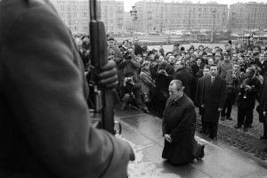 Links im Bildvordergrund ist die Schulter eines Soldaten mit Gewehr zu erkennen. Im Bildzentrum kniet Willy Brandt, er hält die Hände verschränkt vor sich und den Blick nach unten gerichtet. Hinter ihm befindet sich eine Menschenmenge aus Journalisten und Schaulustigen.