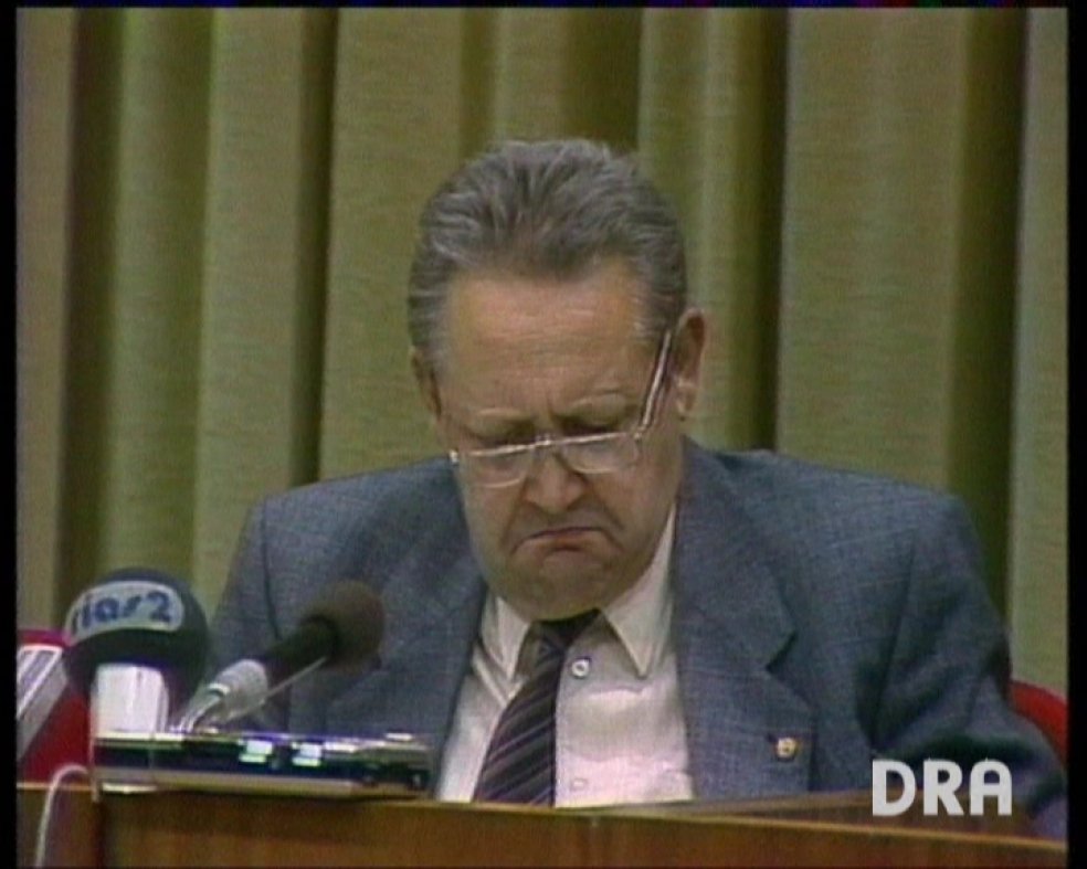Günter Schabowski sitzt vor Mikrofonen und schaut mit ärgerlichem Blick vor sich auf den Tisch.