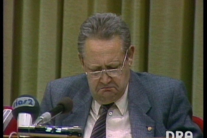Günter Schabowski sitzt vor Mikrofonen und schaut mit ärgerlichem Blick vor sich auf den Tisch.