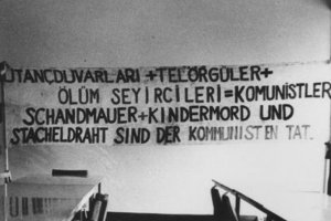 An der Wand hängt ein Banner mit der Aufschrift in deutsch und türkisch: Schandmauer und Kindermord und Stacheldraht sind der Kommunisten Tat. Im Vordergrund stehen Tischreihen und Stühle.