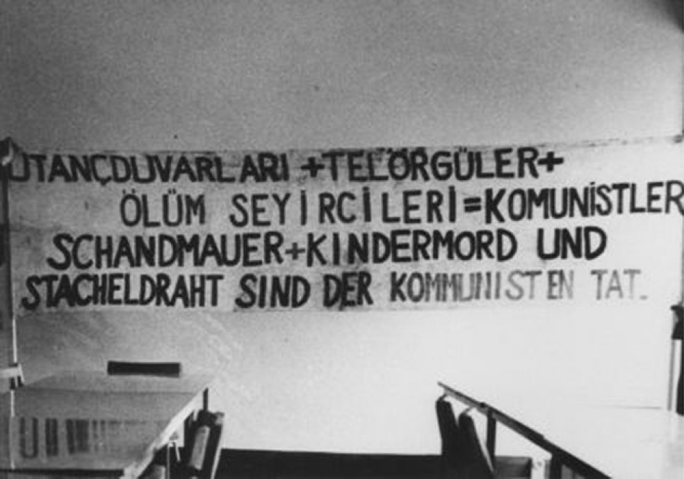 An der Wand hängt ein Banner mit der Aufschrift in deutsch und türkisch: Schandmauer und Kindermord und Stacheldraht sind der Kommunisten Tat. Im Vordergrund stehen Tischreihen und Stühle.