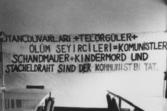 An der Wand hängt ein Banner mit der Aufschrift in deutsch und türkisch: Schandmauer und Kindermord und Stacheldraht sind der Kommunisten Tat. Im Vordergrund stehen Tischreihen und Stühle.