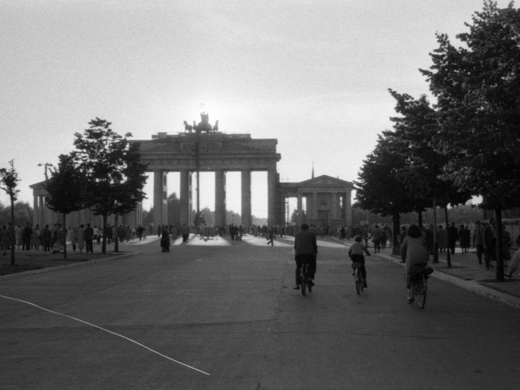 Das Brandenburger von der Straße Unter den Linden in Richtung Tiergarten fotografiert. Dutzende von Menschen sind zu Fuß oder auf dem Fahrrad unterwegs.