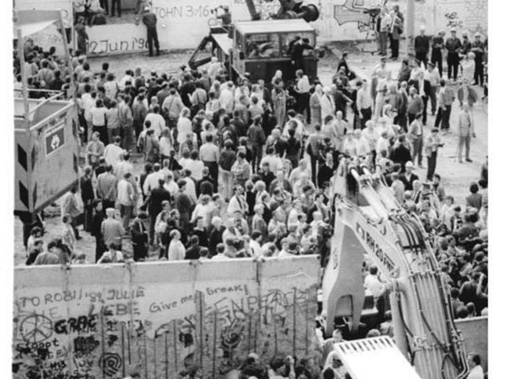 Das Bild zeigt die Grenzanlagen in der Ackerstraße aus der Vogelperspektive. Zentral ist der Blick in den Todesstreifen zwischen der Außenmauer und der Hinterlandmauer. Sowohl außerhalb als auch zwischen beiden Mauern stehen Menschenansammlungen. Zwei Bagger sind dabei, die Mauern abzureißen.