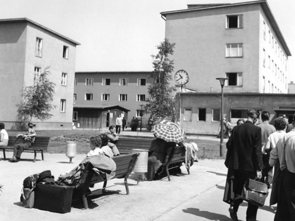 Im Hintergrund ist ein mehrgeschossiges Gebäude zu sehen, auf dem Platz davor sitzen Menschen auf Bänken oder gehen in Richtung des Gebäudes. Einige haben Gepäck dabei.
