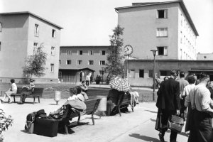 Im Hintergrund ist ein mehrgeschossiges Gebäude zu sehen, auf dem Platz davor sitzen Menschen auf Bänken oder gehen in Richtung des Gebäudes. Einige haben Gepäck dabei.