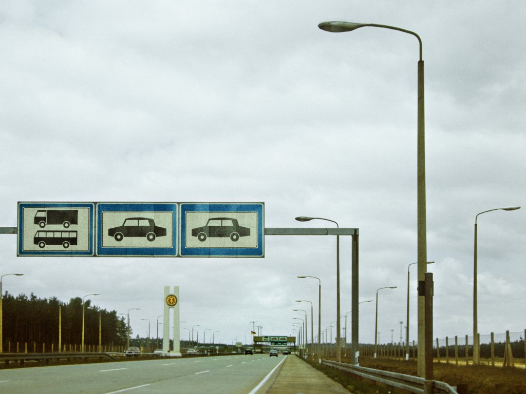 Autobahn A111 mit Blick auf Schilder zur Verkehrsregelung, Lampenmasten und Hoheitszeichen der DDR des Grenzübergangs Stolpe; Aufnahme 15. April 1990