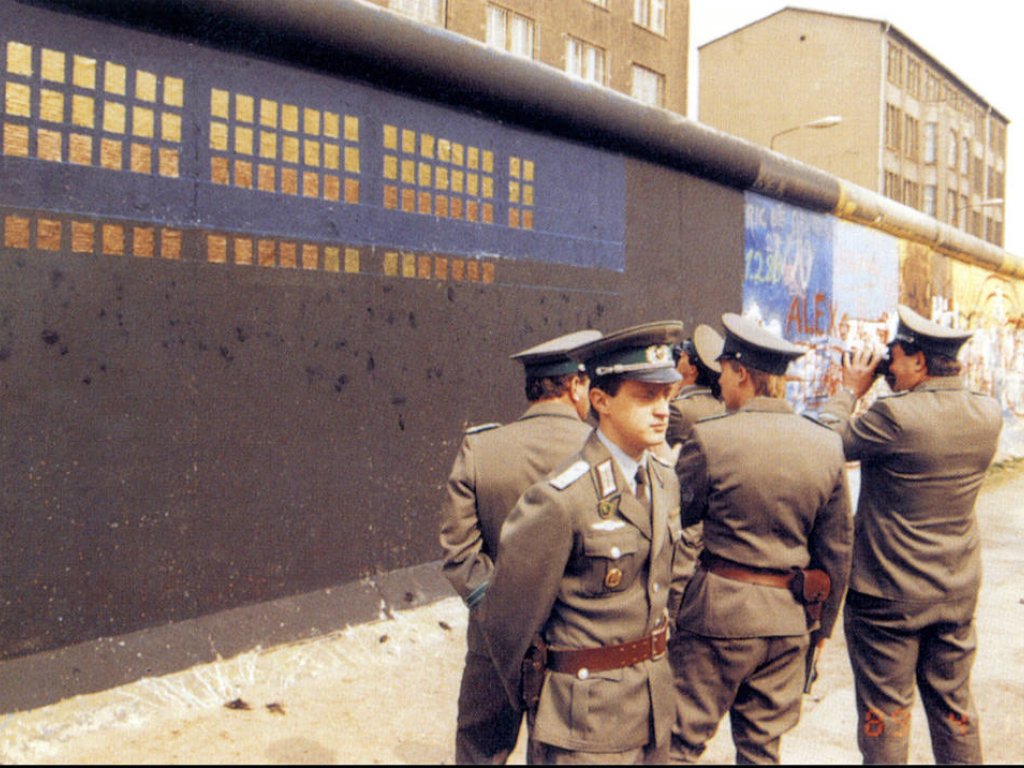 Vor der schwarz-blau grundierten, teilweise mit Kupferplatten beklebten Mauer stehen fünf Grenzsoldaten. Einer filmt die Installation mit einer Kamera. Im Hintergrund sind weitere bemalte Mauersegmente zu sehen.