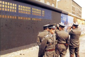 Vor der schwarz-blau grundierten, teilweise mit Kupferplatten beklebten Mauer stehen fünf Grenzsoldaten. Einer filmt die Installation mit einer Kamera. Im Hintergrund sind weitere bemalte Mauersegmente zu sehen.