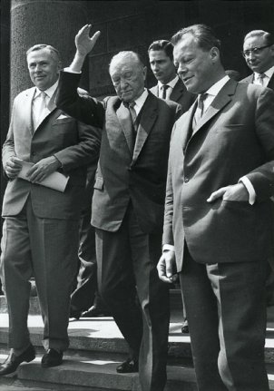 Willy Brandt geht rechts im Vordergrund eine Treppe hinunter. Neben ihm wirft Adenauer den Arm gestikulierend in die Luft. Daneben und dahinter gehen weitere Männer.