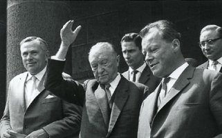 Willy Brandt geht rechts im Vordergrund eine Treppe hinunter. Neben ihm wirft Adenauer den Arm gestikulierend in die Luft. Daneben und dahinter gehen weitere Männer.
