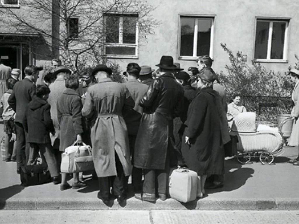 Eine Menschenansammlung steht vor einem Gebäude. Die Rücken sind der Kamera zugewandt, einige haben Gepäck dabei. Rechts ist ein Kinderwagen zu sehen.