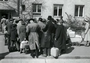 Eine Menschenansammlung steht vor einem Gebäude. Die Rücken sind der Kamera zugewandt, einige haben Gepäck dabei. Rechts ist ein Kinderwagen zu sehen.