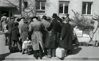 Eine Menschenansammlung steht vor einem Gebäude. Die Rücken sind der Kamera zugewandt, einige haben Gepäck dabei. Rechts ist ein Kinderwagen zu sehen.