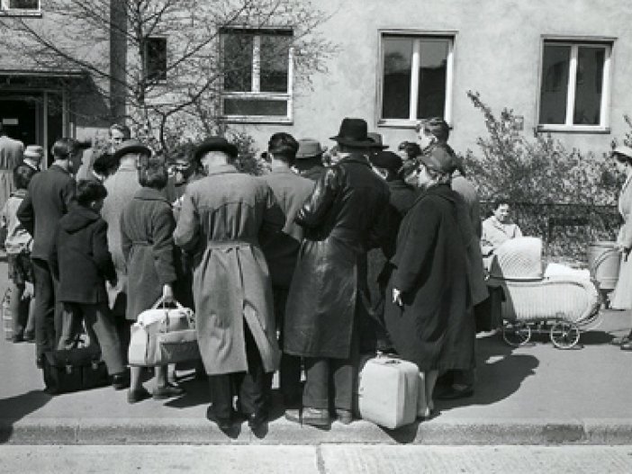Eine Menschenansammlung steht vor einem Gebäude. Die Rücken sind der Kamera zugewandt, einige haben Gepäck dabei. Rechts ist ein Kinderwagen zu sehen.