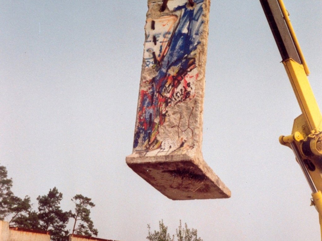 Geschenk der DDR-Regierung an die John F. Kennedy Library in Boston/USA: die bunte Westseite des Mauersegments 8A, August 1990 Ein mit Graffiti besprühtes Mauersegment wird mit einem Kran in die Luft gehoben. Am Boden stehen weitere einzelne bemalte Segmente nebeneinander.