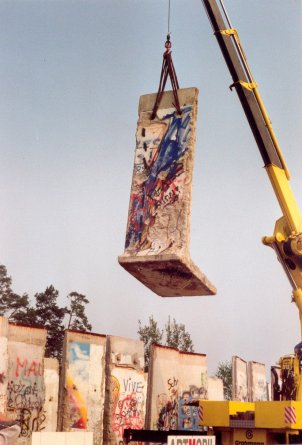 Ein mit Graffiti besprühtes Mauersegment wird mit einem Kran in die Luft gehoben. Am Boden stehen weitere einzelne bemalte Segmente nebeneinander.