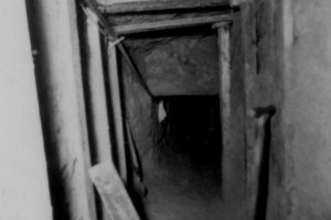 Das Bild zeigt in den Tunnelschacht, der an Decke und Wänden mit Holzbalken abgestützt ist. Im Vordergrund lehnt ein Spaten an der Wand.