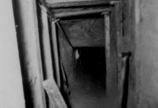 Das Bild zeigt in den Tunnelschacht, der an Decke und Wänden mit Holzbalken abgestützt ist. Im Vordergrund lehnt ein Spaten an der Wand.