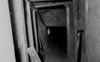 Das Bild zeigt in den Tunnelschacht, der an Decke und Wänden mit Holzbalken abgestützt ist. Im Vordergrund lehnt ein Spaten an der Wand.