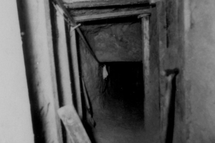 Das Bild zeigt in den Tunnelschacht, der an Decke und Wänden mit Holzbalken abgestützt ist. Im Vordergrund lehnt ein Spaten an der Wand.