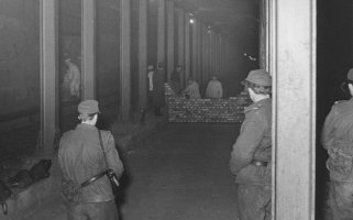 Das Bild zeigt einen mit Stahlträgern abgestützten Schacht. Im Vordergrund stehen drei Soldaten mit dem Rücken zur Kamera und blicken in Richtung einer Mauer aus Ziegelsteinen, die von mehreren Maurern zwischen den Stahlträgern hochgezogen wird.